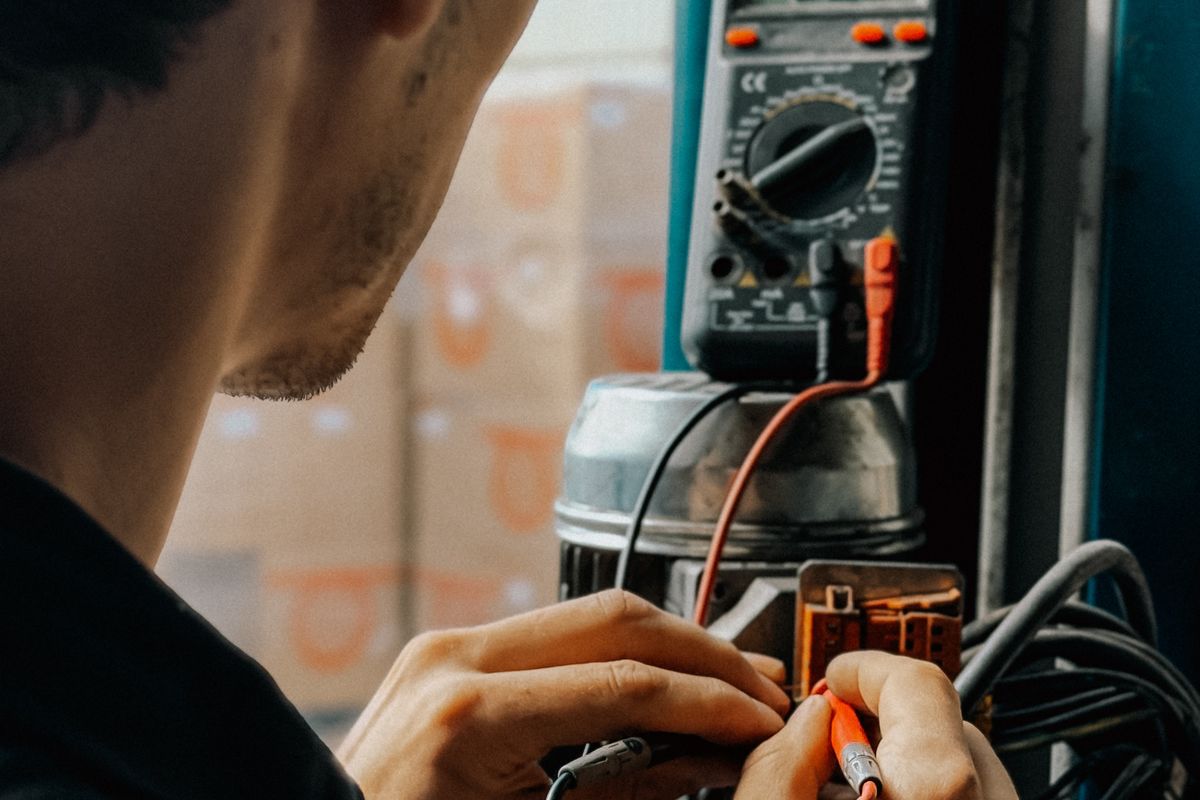 Technicien penché sur un moteur électrique ouvert, pointes de touche d'un multimètre à la main, devant des racks de dépôt orange